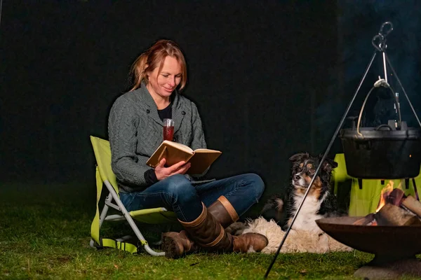 Mujer joven y su pastor australiano afuera por una fogata. Leyendo un libro al anochecer. Pan, queso y vino en la mesa. Caldero cuelga sobre el fuego en invierno — Foto de Stock