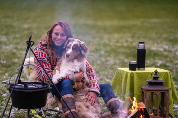 Tricolor pastor australiano se sienta al lado de una joven junto a la fogata. En invierno, nieve en la hierba. El fuego está ardiendo bajo el caldero. Enfoque selectivo, piel, mesa, café en termo — Foto de Stock