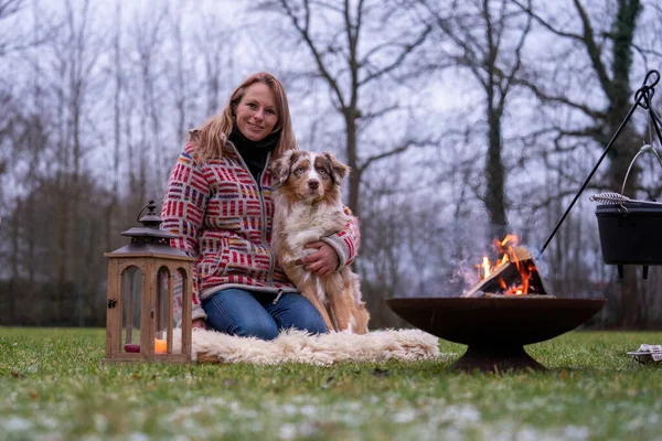 Tricolor pastor australiano se sienta al lado de una joven junto a la fogata. En invierno, nieve en la hierba. El fuego arde al atardecer. Abrazar al perro. Enfoque selectivo — Foto de Stock