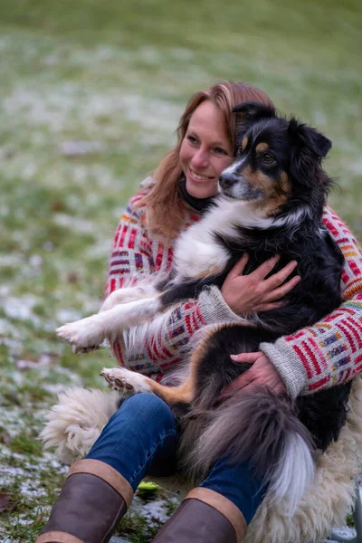 Sarışın kadın, kucağında Avustralya çoban köpeği var. Kışın kamp ateşinin yanında, karlı çimlerin üzerinde, günlük, kalın bir kazakla.