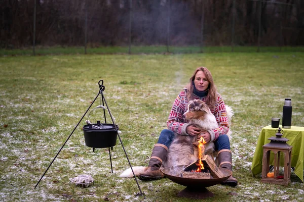 Австралийская овчарка Триколор сидит рядом с молодой женщиной у костра. Зимой снег на траве. Огонь горит под котлом. Селективный фокус, мех, стол, кофе в термосе — стоковое фото