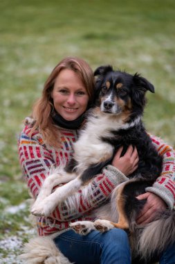 Sarışın kadın, kucağında Avustralya çoban köpeği var. Günlük giysiler, kışlık botlar ve kalın kışlık süveter ile kışın kamp ateşinin yanındaki çimlerin üzerinde.