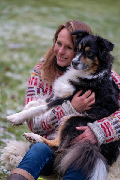 Mujer rubia con su perro pastor australiano en su regazo. En suéter casual de invierno grueso Fuera en la hierba nevada junto a la fogata en invierno — Foto de Stock