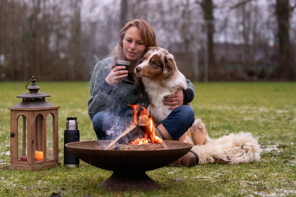 El perro se sienta junto al fuego con los anfitriones y HuskyAustralian Shepherd se sienta al lado de su dueño junto al fuego. La mujer bebe café de una taza de termo. Juntos se sientan en una alfombra de piel en la nieve cubierta — Foto de Stock