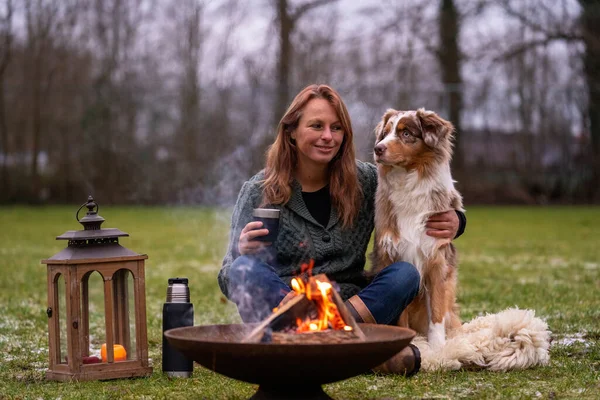 El perro se sienta junto al fuego con los anfitriones y HuskyAustralian Shepherd se sienta al lado de su dueño junto al fuego. La mujer bebe café de una taza de termo. Juntos se sientan en una alfombra de piel en la nieve cubierta — Foto de Stock