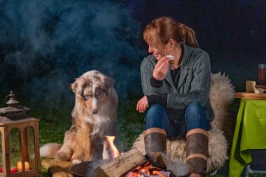Genç bayan peynirli bir dilim baget yiyor. Genç Avustralyalı çoban köpeği yanına oturmuş yavru mavi gözleriyle kamp ateşine bakıyor. Bir kış akşamında kamp ateşinden çıkan duman.