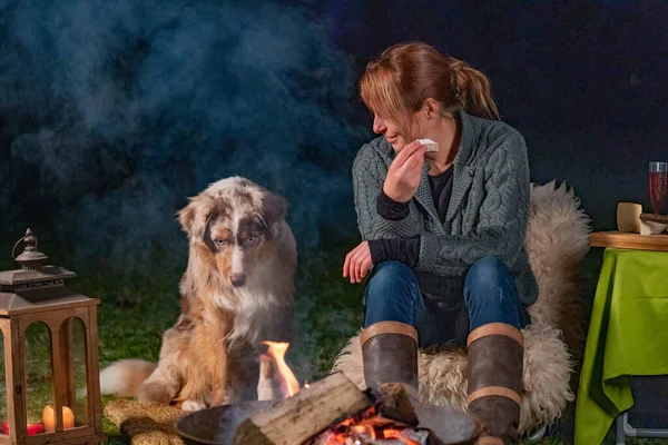 Молодая женщина ест кусочек багета с бри. Её молодая австралийская овчарка сидит рядом с ней, глядя на костёр своими щенячьими голубыми глазами. Дым от костра в зимний вечер — стоковое фото