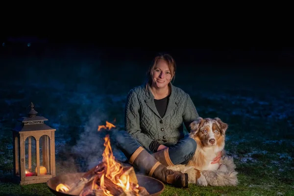 El perro se sienta junto a la fogata con su dueño. La mujer mira con amor al pastor alemán. Vacaciones en invierno, fuego de vela en la linterna — Foto de Stock