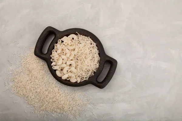 Gluten-free italian pasta products (pipe rigate, vermicelli) made from rice flour in wooden serving platter. Top view, grey textured background, copy space.