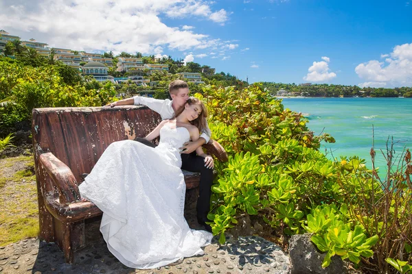 Happy bride and groom sitting on the bench in front of beautiful — Stock Photo, Image