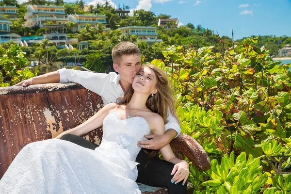 Happy bride and groom sitting on the bench in front of beautiful — Stock Photo, Image