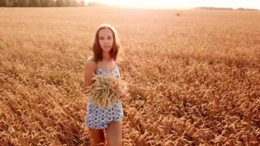 Genç ve güzel bir kız gün batımında altın olgun buğday tarlasında yürüyor..