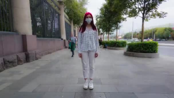 Проміжок часу дівчинки-підлітка в червоному волоссі в масці covid19 стоїть в центрі зайнятої вулиці, дивлячись на камеру, в той час як юрби людей йдуть повз. Вірус інфекції пандемії грипу корона вірус 2019-ncov. — стокове відео