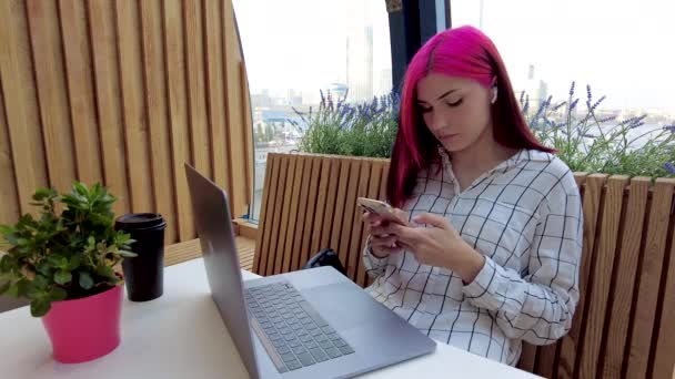 Belle étudiante rousse travaille comme employée à distance avec ordinateur portable et smartphone dans un café. — Video