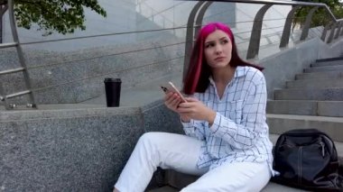 Üzgün, kızıl saçlı, akıllı telefonlu öğrenci şehirde merdivenlerde oturan sosyal ağlarda sıkılıyor..
