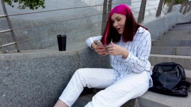 Şehirdeki merdivenlerde oturan güzel kızıl saçlı öğrenci kız sosyal ağlarda iyi haberler almaktan mutlu..