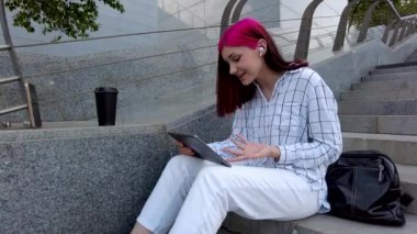 Güzel kızıl saçlı kız arkadaşlarıyla iletişim kuruyor. Sosyal ağlarda, şehirdeki basamaklarda oturan tablet üzerinden..