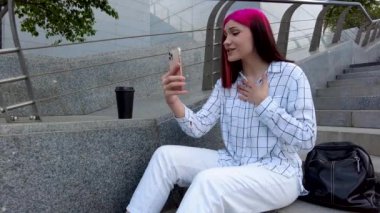 Güzel, kızıl saçlı, etkili blogcu kız, şehirdeki basamaklarda oturan sosyal ağlardaki aboneleriyle video konuşması yapıyor..
