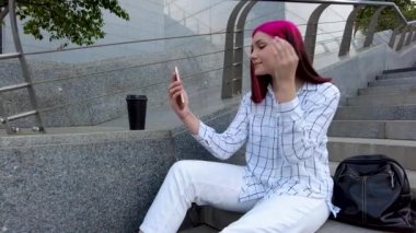 Güzel kızıl saçlı öğrenci poz veriyor. Sosyal ağlar için akıllı telefondan selfie çekiyor. Şehrin basamaklarında oturuyor..