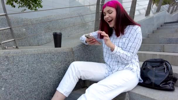 Glückliche schöne rothaarige Studentin lacht herzlich und plaudert in sozialen Netzwerken auf den Stufen der Stadt mit dem Smartphone. — Stockvideo