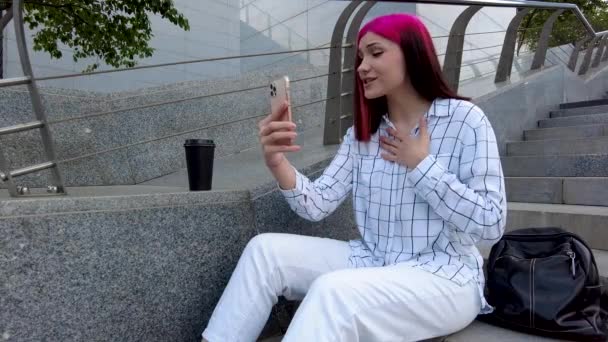 Bela blogueira influenciadora ruiva conversando fazendo videochamadas usando smartphone com seus assinantes nas redes sociais sentados nos degraus da cidade. — Vídeo de Stock