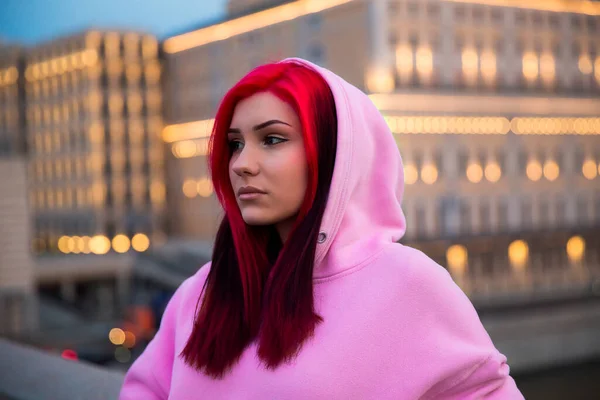 Sad red-haired teenage girl in pink hoodie in the evening on lighted city street
