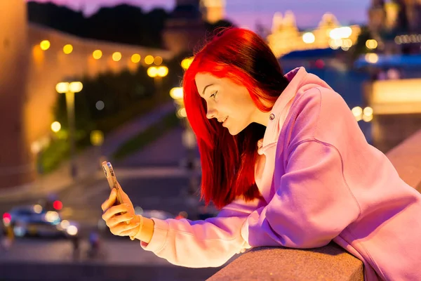 Beautiful red-haired teenage girl in pink hoodie talking using smartphone with her subscribers in social networks in the evening on lighted city street