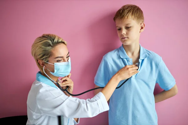 Ung kvinnlig läkare i kirurgisk mask med hjälp av ett stetoskop för att kontrollera pulsen — Stockfoto