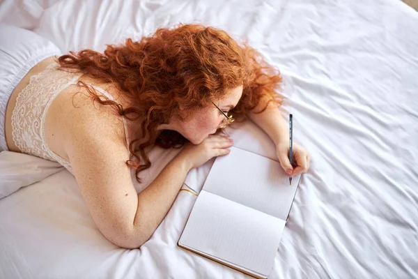 Bonita pelirroja semidesnuda hacer notas tumbado en la cama —  Fotos de Stock