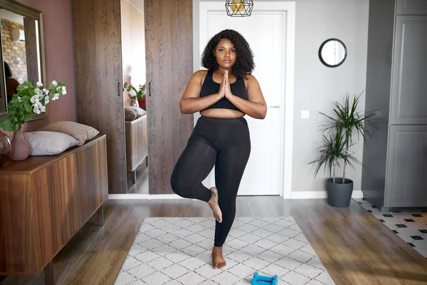 Tyk sort kvinde holde balancen, stå på det ene ben, yoga tid hjemme - Stock-foto