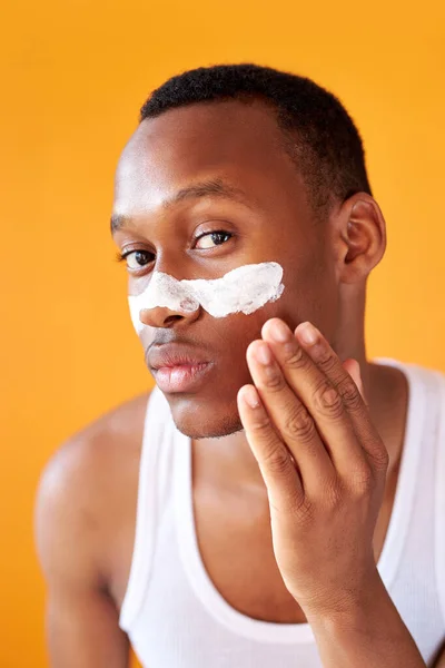 Fiatal afro-amerikai srác alkalmazása arc krém a szeme alatt sárga háttér — Stock Fotó