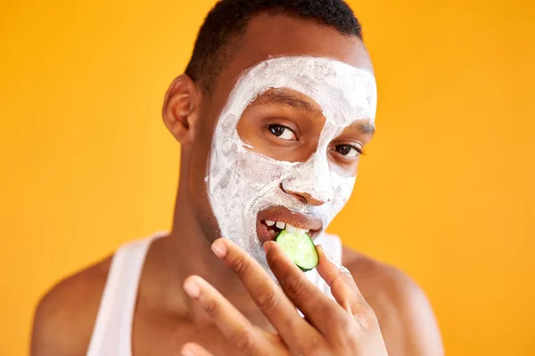 Retrato de hombre negro loco comiendo pepino mientras tiene máscara de belleza facial —  Fotos de Stock