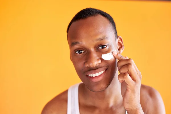 Jeune homme afro-américain appliquant de la crème visage sous ses yeux sur fond jaune — Photo