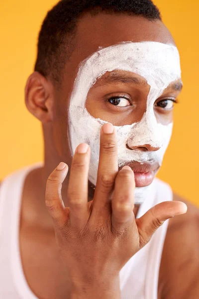 Ung afroamerikansk fyr som bruker ansiktskrem under øynene på gul bakgrunn – stockfoto