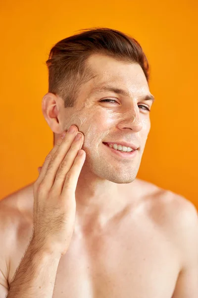Νεαρός καυκάσιος άντρας εφαρμόζει ενυδατική κρέμα προσώπου για υγιές λαμπερό δέρμα, απομονωμένο — Φωτογραφία Αρχείου