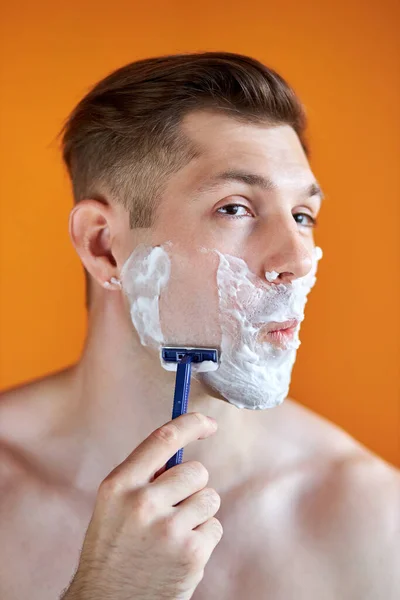 Portrait of caucasian male shaving his face with foam and razor isolated on orange background — Stock Photo, Image