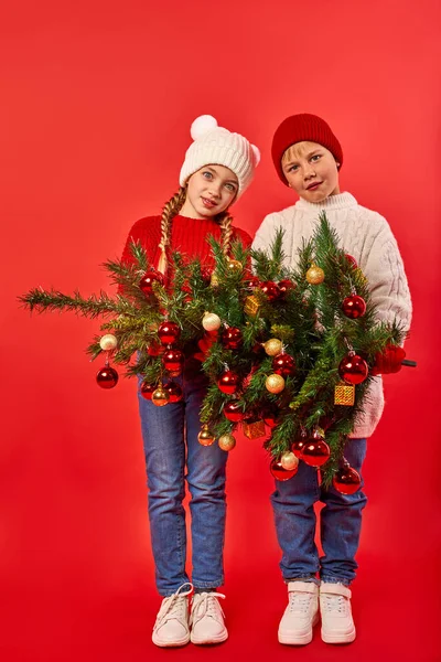 Noel ağacını ellerinde tutan iki sevimli çocuğun tam boy portresi.