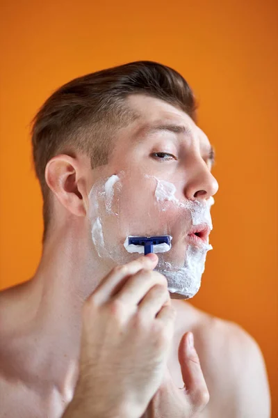 Retrato de hombre caucásico afeitándose la cara con espuma y navaja aislada sobre fondo naranja — Foto de Stock