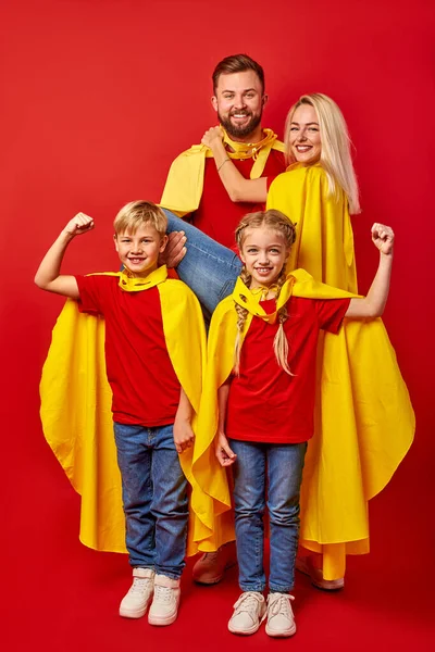 Family of superheroes in costumes posing in studio with red background — Stock Photo, Image