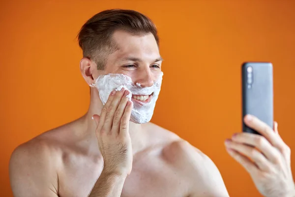 smiling man cheerfully apply shaving foam and looking at camera of smartphone