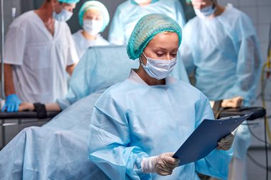 Kadın hemşire, meslektaşları ameliyat yaparken dijital tablet üzerine bir belgeyi okumaya odaklanmıştır.