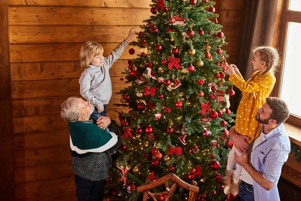 elderly man and young male hold children in hands, they decorate christmas tree