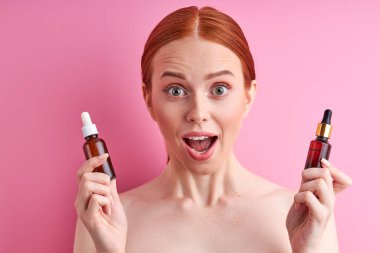 Heyecanlı kızıl saçlı üstsüz kadın elinde iki şişe serum tutuyor.