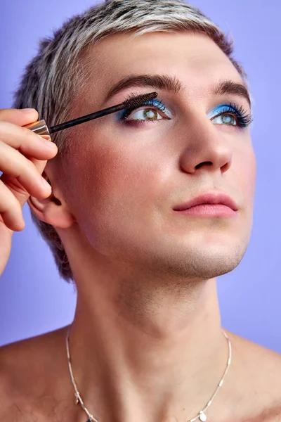 Ung transgender man tillämpa mascara på ögonfransar mot färg bakgrund — Stockfoto