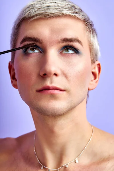 Πορτρέτο του νεαρού, όμορφου ομοφυλόφιλου άνδρα που βάζει μακιγιάζ, μάσκαρα βλεφαρίδων απομονωμένη — Φωτογραφία Αρχείου