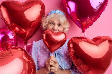 Şok olmuş olgun bir kadın parti veriyor, kırmızı balonlar takıyor, şık kıyafetler giyiyor.