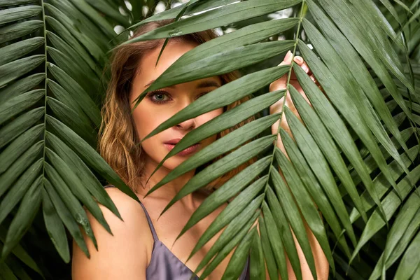 Close-up retrato de jovem mulher confiantemente olhando para a câmera através de folha de planta exótica — Fotografia de Stock