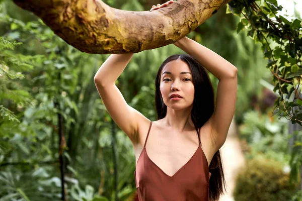 Розкішна брюнетка в елегантній шовковій сукні позує в тропічному саду біля дерева — стокове фото