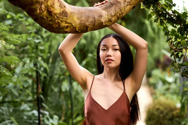 Linda mulher morena em vestido de seda elegante posando no jardim tropical perto da árvore — Fotografia de Stock