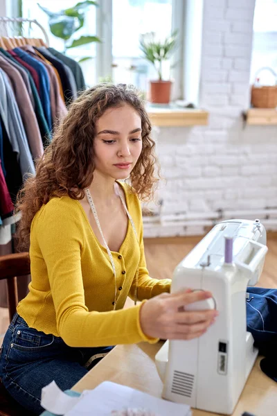 attractive caucasian dressmaker tailoring while sitting at the sewing machine looking seriously and confident